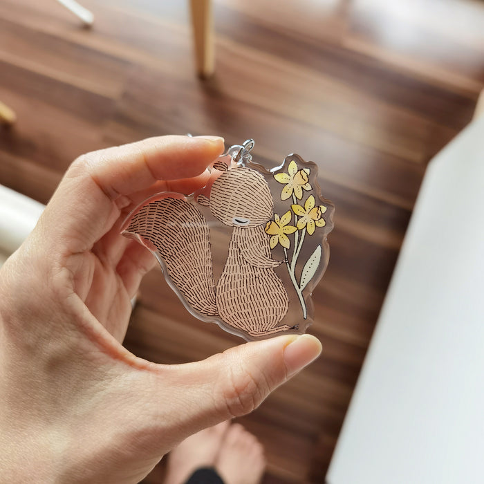 Acrylic keycharm featuring a squirrel sniffing daffodil, adds nature&#39;s beauty to bags and keys, a stylish accessory.