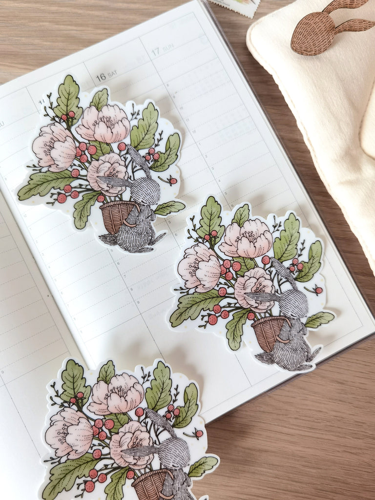 Waterproof sticker featuring rabbit carries a basket full of blooms. Add a touch of floral charm to laptops, journals, sketchbooks, and any surface.