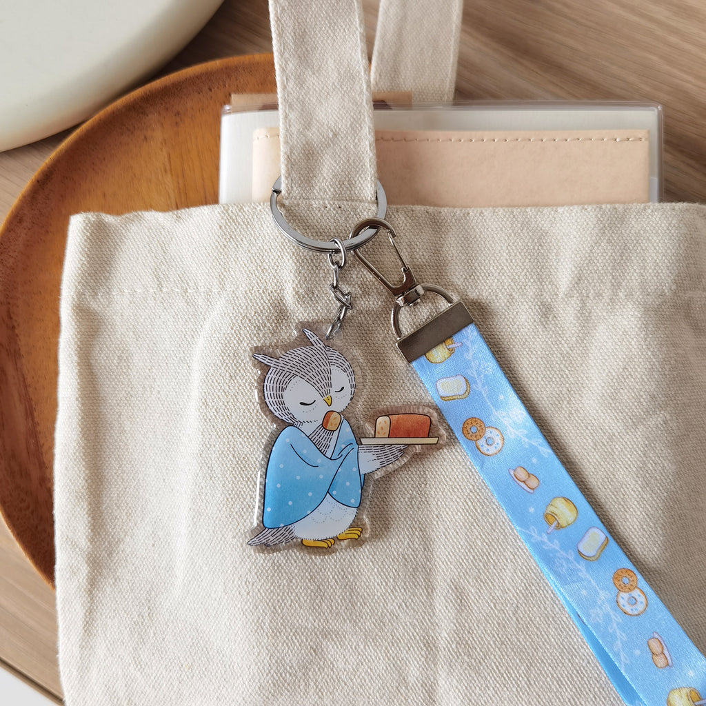 Cute owl savoring orange cake key charm with matching lanyard of illustrated honey toast, scones, and bagel. A whimsical accessory for bags or keys.