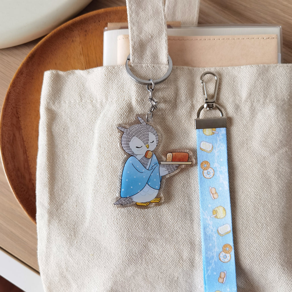 Cute owl savoring orange cake key charm with matching lanyard of illustrated honey toast, scones, and bagel. A whimsical accessory for bags or keys.