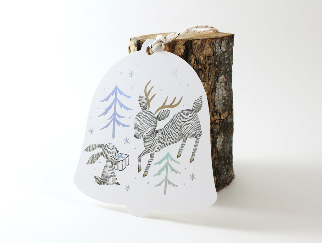 Holographic Foil Christmas Holiday Gift Tags - Rabbit, Reindeer & Christmas Gift - winter forest trees elegantly printed in holographic foil, whimsical illustration of rabbit presenting a gift to a reindeer, custom die-cut in the shape of a bell, adorned with cotton twine.