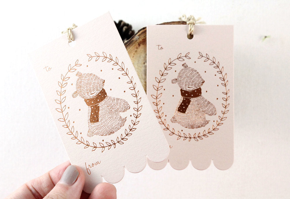Rectangular Christmas gift tag with illustrated winter bear and festive wreath design in shimmering copper foil on blush pink cardstock. Scalloped edge on one side. Ideal for holiday gifting and Christmas presents.