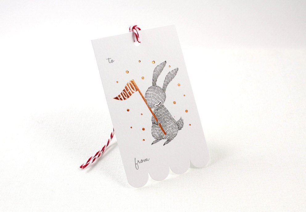 Rectangular Christmas gift tag with illustrated rabbit holding a copper foil "HOHO" party flag, on sturdy cardstock. Scalloped edge on one side. Ideal for holiday gifting.