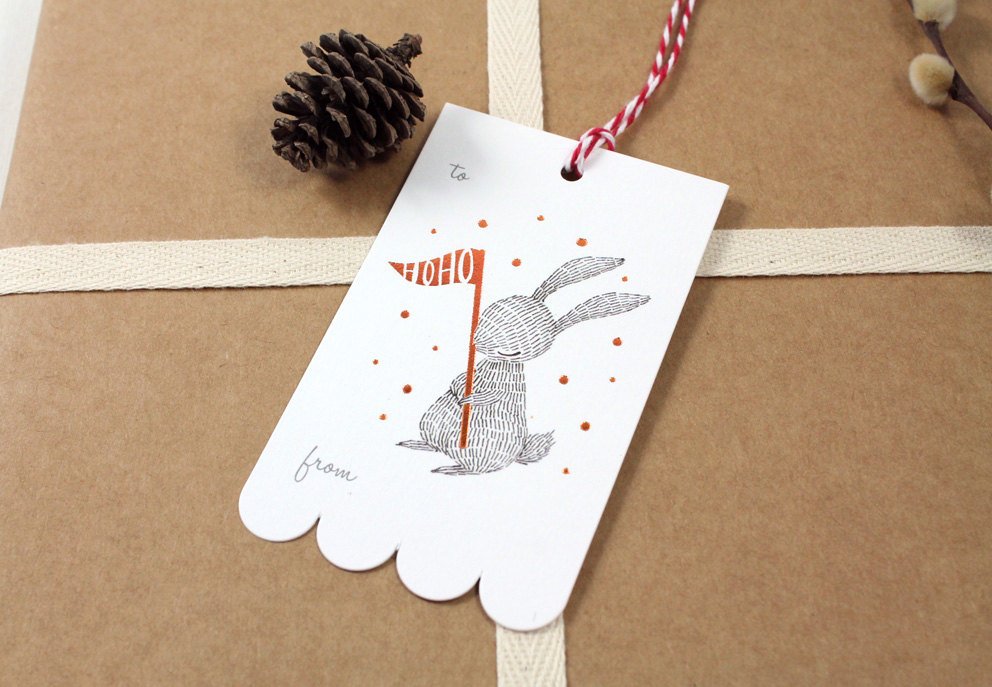Rectangular Christmas gift tag with illustrated rabbit holding a copper foil "HOHO" party flag, on sturdy cardstock. Scalloped edge on one side. Ideal for holiday gifting.